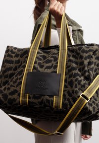 Femme tenant un grand sac de voyage en toile à imprimé léopard noir et marron, avec des poignées rayées jaunes et noires et un patch logo en cuir noir.