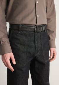 Črne denim hlače z vidnimi šivi in sprednjimi žepi, kombinirane s temno rjavim srajco z umetno črtami in črnim usnjenim pasom.