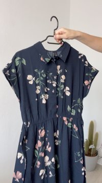 Navy krāsas kleita ar ziedu rakstu rozā un zaļā krāsā, īsām piedurknēm, ar apkakli, elastīga jostasvieta, plūstošs audums, karājas no melna pakaramā.