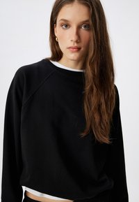 Zwarte cropped sweatshirt met een ronde hals en raglanmouwen, met een gestructureerde stof en een gelaagd wit shirt eronder.