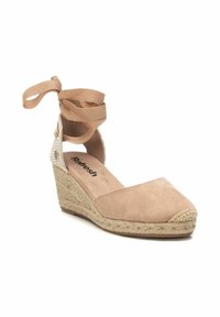 Espadrille de cuña beige con una suela de yute texturizada, parte superior de ante y tiras de tela en el tobillo atadas en un lazo. Presenta una puntera redondeada y una plantilla acolchada.