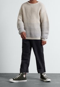 Bambino che indossa un maglione lavorato a maglia beige e grigio, pantaloni neri arrotolati e sneaker alte verdi, in piedi su un pavimento grigio.