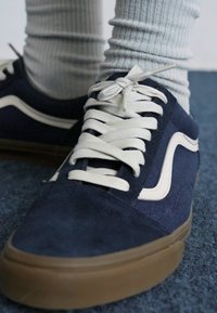 Marineblå semsket sneakers med beige detaljer, hvite snører og gummisåle. Ribbestrikkede lysegrå strømper synlige over skoen.