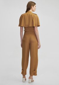 Korte bruine top met cape-mouwen, gecombineerd met hooggelegde, wijde broeken met knoopmanchetten en een gladde textuur.