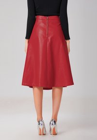 Jupe midi en cuir rouge avec un design évasé, fermeture éclair au dos et texture lisse. Associée à des chaussures à talons hauts argentées.