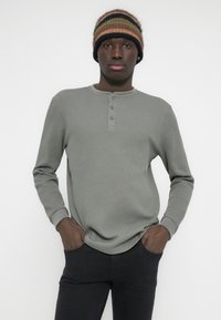 Maglietta a maniche lunghe in maglia waffle grigia con tre bottoni al collo, indossata con pantaloni neri aderenti. Modella posa con le mani nelle tasche.