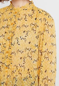 Camicia gialla trasparente con collo arricciato, bottoni sul davanti e un motivo floreale in marrone e nero. Tessuto leggero.