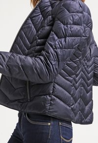 Veste matelassée bleu marine avec un motif chevrons matelassés, fermeture éclair et silhouette courte. Le matériau semble lisse et brillant.