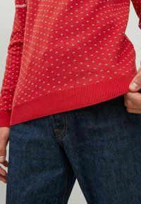 Roter Strickpullover mit einem strukturierten Muster aus kleinen weißen Punkten, mit einem gerippten Saum, kombiniert mit dunkelblauen Denim-Jeans.