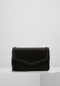 Bolso de cuero sintético negro con una correa de cadena, que presenta un diseño de sobre y un detalle de borde cosido, colocado sobre una superficie blanca.