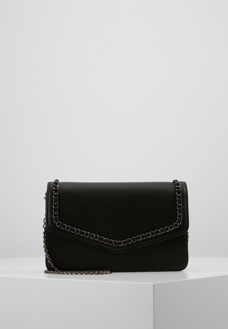 Bolso de cuero sintético negro con una correa de cadena, que presenta un diseño de sobre y un detalle de borde cosido, colocado sobre una superficie blanca.