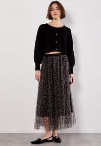 Frau in einem schwarzen, geknöpften, cropped Sweater, einem Tüll-Midi-Rock mit Leopardenmuster und schwarzen flachen Slingback-Schuhen, die gegen eine weiße Wand steht.