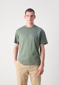 T-shirt en coton vert avec un col rond, des manches courtes et un ourlet légèrement arrondi. Design simple avec un logo discret sur la poitrine.