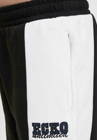 Black and white joggers with an elastic waistband, featuring a side pocket and an embroidered "ECKO" logo in blue on the thigh.