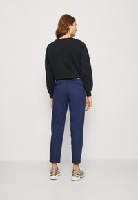 Felpa nera, pantaloni tapered blu scuro, due tasche posteriori e sneakers con motivo. Il tessuto sembra liscio; la scarpa presenta dettagli arancioni.