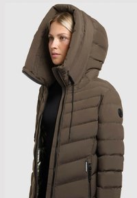 Padded hnedá bunda s veľkou, predĺženou kapucňou, zipsovým zapínaním a bočnými vreckami. Obsahuje prešívaný dizajn a logo nášivku na rukáve.