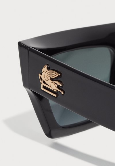 Black sunglasses with a geometric design. Features a shiny gold pegasus emblem on the side and tinted lenses. Curved and angular shapes.