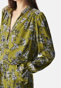 Grüne Blumenbluse mit langen Ärmeln, einem tiefen V-Ausschnitt und kunstvollen schwarz-weißen botanischen Mustern. Hergestellt aus glattem Stoff.