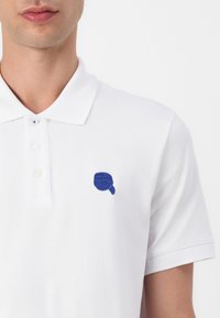 Biely polo tričko z textúrovanej látky, s malým modrým vyšívaným dizajnom na ľavej hrudi a klasickým golierom so zoznamom troch gombíkov.