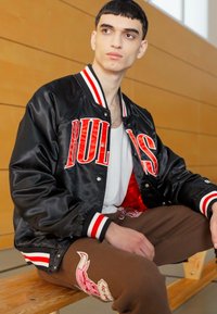 Svart satin bomberjacka med röd "BULLS" text, randiga ribbade manschetter och en vit undertröja. Bruna byxor med en rosa broderad design.