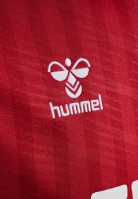 Rød sportskjorte med brodert hvit 'hummel'-logo og et bie-design, med en teksturert stoffoverflate.