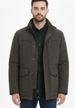 Hombre vestido con una chaqueta de cierre verde oscuro con bolsillos con solapa y bolsillos en el pecho con cremallera sobre una camisa de cuadros verde oscuro, de pie contra un fondo blanco.