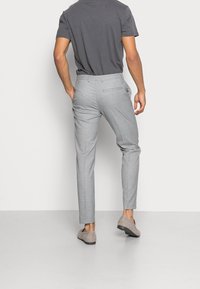Uomo che indossa pantaloni slim-fit grigio chiaro, mocassini grigi in camoscio e una camicia a maniche corte grigio scuro, in piedi davanti a uno sfondo bianco semplice.