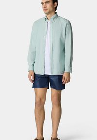 Lichtgroen button-up shirt met lange mouwen, gedragen over een wit T-shirt, gecombineerd met marineblauwe shorts. De stijl is casual en eenvoudig.