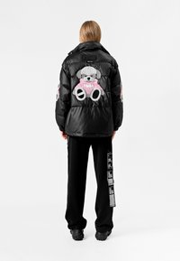 Chaqueta de pluma negra con un diseño de oso de peluche en la parte posterior y acentos rosas. Combinada con pantalones de chándal negros y calzado grueso negro.