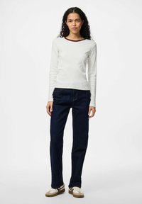 Kvinna med lockigt mörkt hår som bär en vit långärmad topp med vinröd krage, mörkblå jeans och vita och beige sneakers, stående.