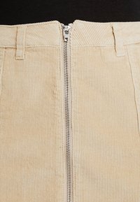 Gonna in corduroy beige con righe verticali, dotata di chiusura con zip frontale e passanti per cintura in vita. Il tessuto ha una texture morbida.