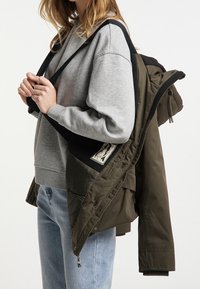 Olivgrön vadderad jacka med svarta detaljer, med dragkedja, huva och logotypmärke. Kombinerad med en grå sweatshirt och ljusa jeans.