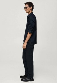 Camicia scura a maniche lunghe e pantaloni abbinati. Occhiali da sole neri eleganti e stivali neri con texture. Design minimalista con una silhouette aderente.