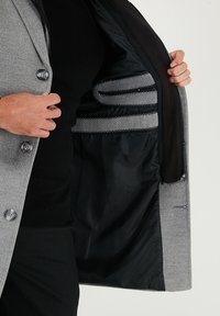 Šedý vlněný kabát s černou saténovou podšívkou, s knoflíky, vnitřními kapsami s ozdobným prošitím a elegantním střihem.