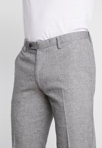 Osoba v šedých kalhotách střední výšky se předním knoflíkem a poutky na opasek, doplněná o bílou košili zastrčenou dovnitř.