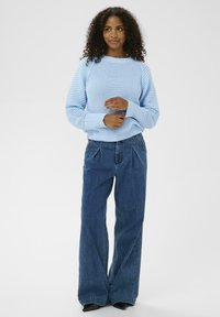 Femme aux cheveux bouclés portant un pull en tricot bleu clair et un jean large bleu foncé, se tenant devant un fond blanc uni.