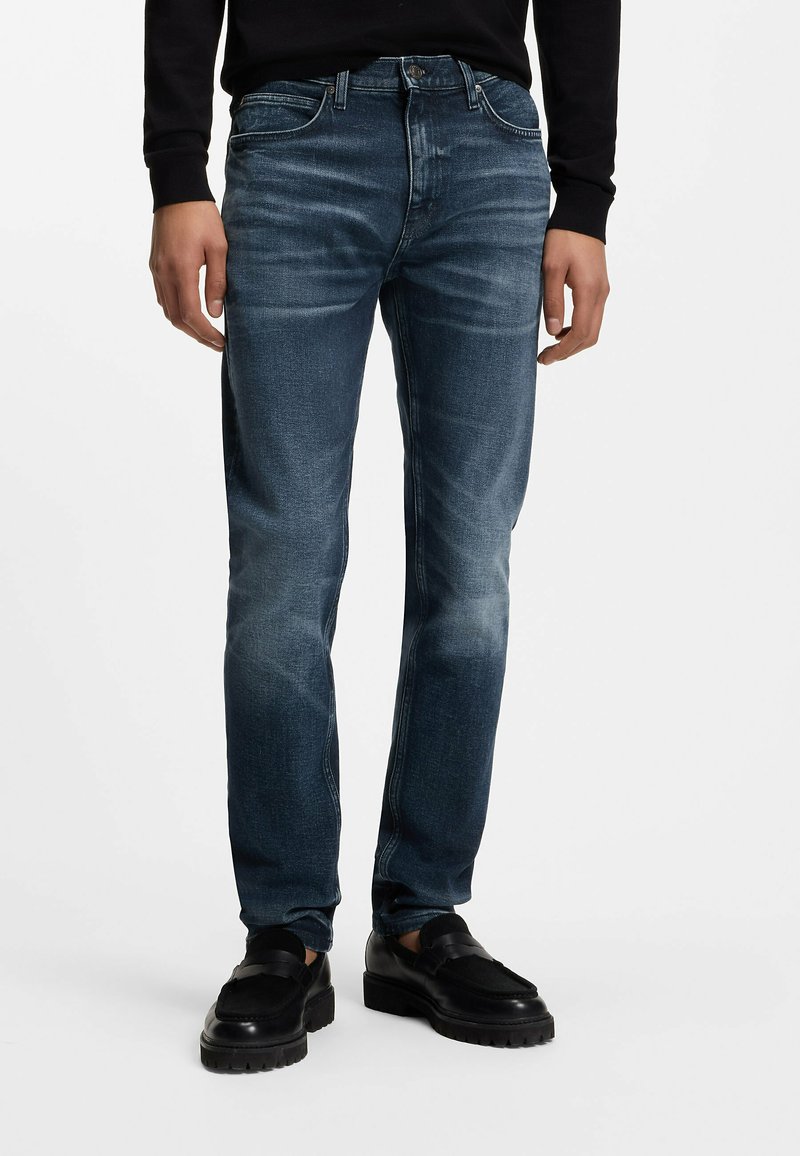 Homme portant un jean bleu délavé et des mocassins noirs, debout devant un fond blanc.