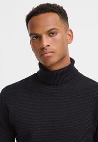 Pulover tricotat bleumarin cu o guler înalt cu nervuri, având un model texturat. Materialul pare gros și călduros, conceput pentru o siluetă ajustată.