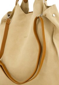 Sac fourre-tout en suède beige avec des poignées en cuir brun, présentant une texture lisse et une fermeture à bouton, mettant en avant un branding minimal à la surface.