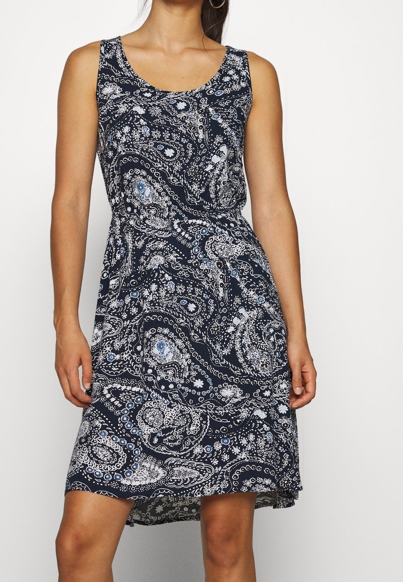 Femme portant une robe bleu marine sans manches, longueur genou, avec un motif floral paisley blanc et bleu clair, debout devant un fond uni.