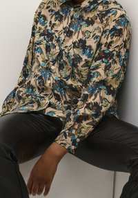 Chemise à boutons florale avec une base beige, présentant des motifs bleus et verts. Portée avec un pantalon en cuir noir, mettant en valeur une coupe décontractée.
