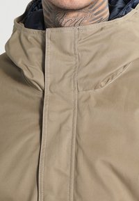 Brązowa kurtka z kapturem o gładkiej fakturze. Posiada wysoki kołnierz, ukryte zapięcie na suwak oraz subtelne detale przeszyć.