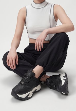 Persona seduta a gambe incrociate che indossa un crop top bianco senza maniche, pantaloni da jogging neri e sneaker chunky nere con suola bianca.
