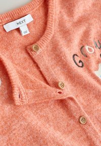 Oranger, gestrickter Cardigan mit Holzknoepfen, einem V-Ausschnitt und dem Schriftzug "Grow" in kontrastierender Stickerei. Weiche Textur ueberall.