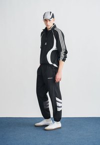 Μαύρο αθλητικό φόρμα με λευκές λεπτομέρειες και τρεις ρίγες στα μανίκια και το παντελόνι. Περιλαμβάνει γκρι καπέλο και λευκά αθλητικά παπούτσια. Ομαλή υφή.