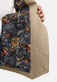 Beige jakke med mønstret marinefarget fôr. Fôret har blomster- og paisleymønstre i rødt, oransje og blått. Merket sier "JEFF Urban Tailoring."