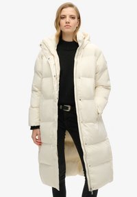 Lang, polstret vinterjakke i off-white. Har hætte, lynlås lukning og trykknapper. Struktureret stof med pænt syede sømme.