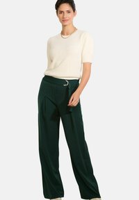 Kremo spalvos megztinis trumpomis rankovėmis, derinamas su aukštu liemeniu tamsiai žaliais plačiomis kelnėmis su diržu, kuriame sidabrinė apvali sagtis.