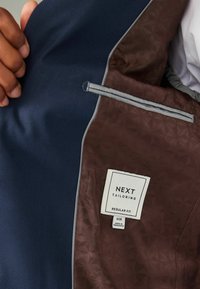 Tumesinine ülikonnajakk, mille sisemise vooder on pruun geomeetrilise mustriga. Omab halli ääristust ja sildit "NEXT Tailoring". Tavalise lõike suurus 40R.