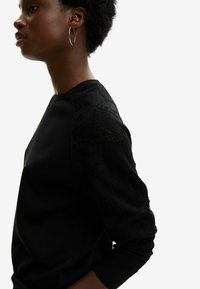 Felpa nera con maniche in maglia texturizzata, scollo rotondo e polsini a coste. Tessuto leggero, silhouette aderente. Orecchini a cerchio argento.